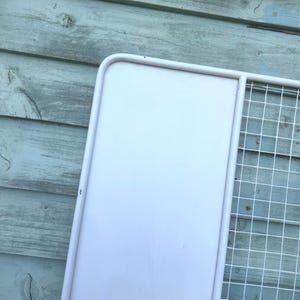 Puede incluir: Una estanter&iacute;a de pared de metal blanco con una parte trasera s&oacute;lida y una secci&oacute;n de rejilla de alambre. La estanter&iacute;a est&aacute; pintada de blanco y tiene un marco redondeado. El fondo es una superficie de madera azul claro desgastada.