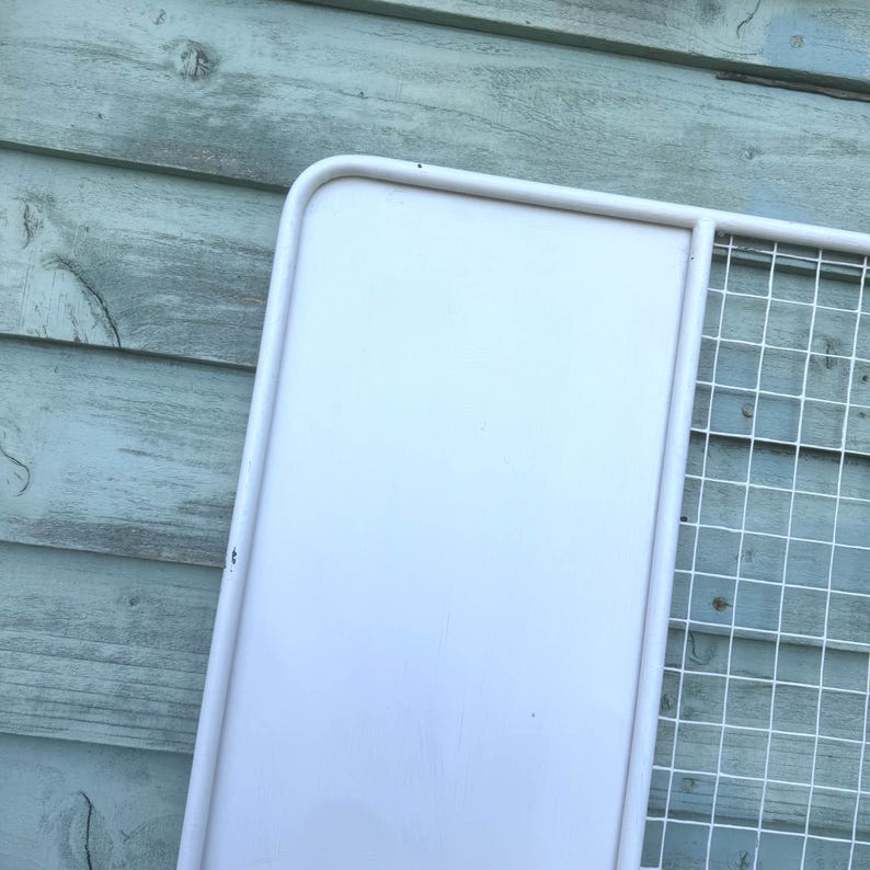 Puede incluir: Una estanter&iacute;a de pared de metal blanco con una parte trasera s&oacute;lida y una secci&oacute;n de rejilla de alambre. La estanter&iacute;a est&aacute; pintada de blanco y tiene un marco redondeado. El fondo es una superficie de madera azul claro desgastada.