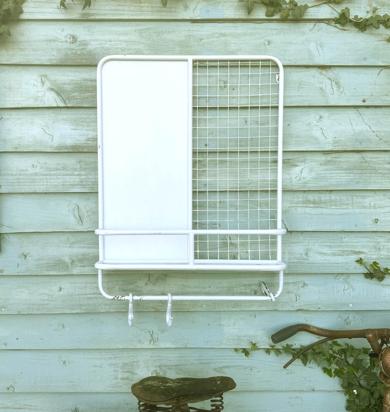 Puede incluir: Organizador de pared de metal blanco con un panel s&oacute;lido y una secci&oacute;n de rejilla. Cuenta con un estante, una barra para toallas y dos ganchos. Montado en una pared de madera azul claro.