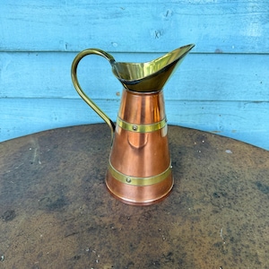 May include: A copper jug with a brass handle and spout. The jug has a cylindrical body with brass bands and rivets. The handle curves upwards, and the spout is wide and angled. The jug is set against a blue wooden background.