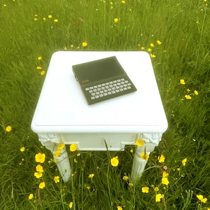 May include: A black ZX81 computer with a QWERTY keyboard and a dark screen sits on a square white table. The device features a compact design with white lettering on the keys and the ZX81 logo printed in orange on the top left corner of the casing.