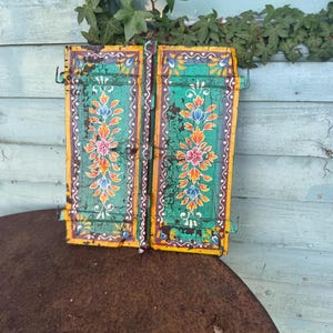 May include: A pair of weathered, rectangular wooden shutters featuring a distressed teal green finish with hand-painted floral motifs in orange, pink, and blue. The edges are trimmed in yellow, and the shutters include metal hinges and hooks.
