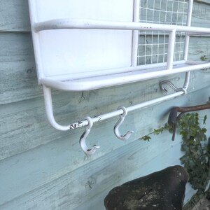 Puede incluir: Estanter&iacute;a de pared de metal blanco con una secci&oacute;n de malla met&aacute;lica y una barra inferior con tres ganchos. La estanter&iacute;a tiene un aspecto envejecido y vintage, con desgaste visible en la pintura. Dise&ntilde;o sencillo y funcional.