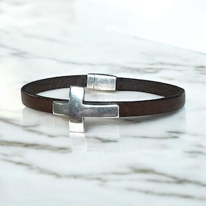 May include: A brown leather bracelet with a silver cross charm. The bracelet has a magnetic clasp. The cross is a prominent feature, adding a religious or symbolic element to the accessory. The bracelet is displayed on a marble surface.