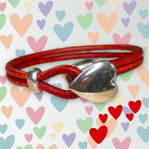 May include: A red leather bracelet with a silver heart-shaped clasp. The bracelet is on a background of colorful hearts.