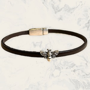 May include: A dark brown leather bracelet with a silver bee charm. The bracelet has a magnetic clasp. The bee charm is detailed with wings and body segments. The bracelet is a simple, elegant accessory.
