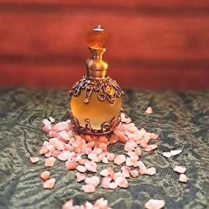 May include: A small, round, frosted glass perfume bottle with a metallic gold-toned filigree overlay featuring amber-colored crystals. The bottle has a matching gold-toned stopper topped with a faceted amber-colored gem, surrounded by small pink stones.