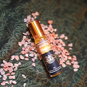 May include: A small glass bottle of "Alchemy Oil Apollo" with a gold cap, resting on a bed of pink crystals. The label is black and gold with white text. The background is a dark green fabric.
