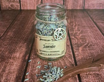 May include: A clear glass jar filled with dried lavender buds, featuring a beige label that reads Apothecary Flowers, Lavender, Lavandula x Intermedia, Woodland Witchcraft. A silver pentagram charm hangs from a twine string around the jar neck.