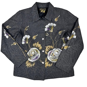 Puede incluir: Chaqueta gris oscuro con diseño floral. La chaqueta presenta flores y hojas pintadas en blanco, morado y dorado. Tiene cierre de botones en la parte delantera y cuello. La chaqueta es de talla mediana.