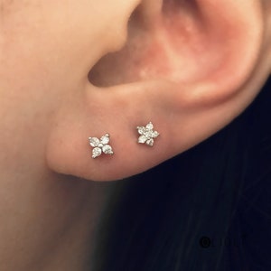 May include: Two small, flower-shaped stud earrings with clear, sparkling stones are shown on an ear. One earring has four stones, and the other has six stones. The earrings are set in a silver-colored metal.