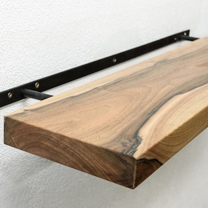 Könnte beinhalten: Ein Holzregal mit einer lebenden Kante und schwarzen Metallkonsolen. Das Regal ist an einer weißen Wand montiert.