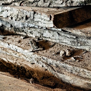 May include: A stack of rough-hewn wooden planks, showing the natural grain and texture of the wood. The planks are weathered and have a rustic appearance.