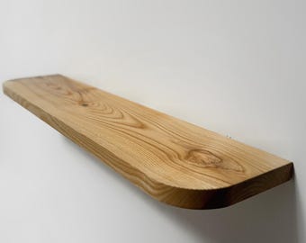 May include: A wall-mounted floating shelf made of natural light-colored wood with a visible grain pattern and a live edge. The shelf features a smooth, polished surface with a prominent knot on the right side and rounded corners on the front edge.