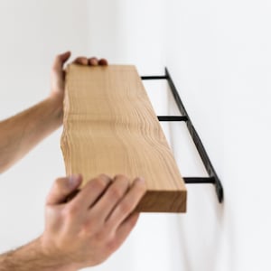 Op de afbeelding: Een persoon die een houten plank met zwarte metalen beugels installeert. De plank is gemaakt van natuurlijk hout met een levende rand en wordt op zijn plaats gehouden door de handen van de persoon.