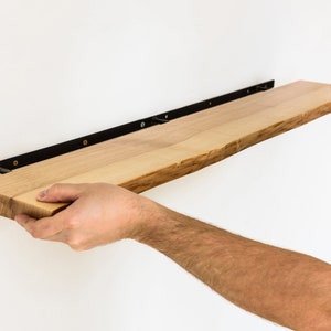 May include: A person installing a wooden shelf with black metal brackets. The shelf is made of natural wood with a live edge and is being secured to a white wall.