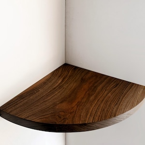 May include: A dark brown wooden corner shelf with a rounded edge. The shelf is mounted on a white wall.