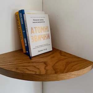 May include: A wooden corner shelf with three books stacked on top. The top book is titled "Atomic Habits: An Easy & Proven Way to Build Good Habits & Break Bad Ones" by James Clear. The other two books are in a foreign language.