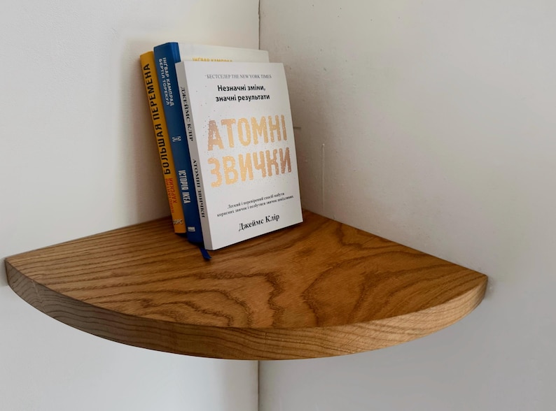 May include: A wooden corner shelf with three books stacked on top. The top book is titled "Atomic Habits: An Easy & Proven Way to Build Good Habits & Break Bad Ones" by James Clear. The other two books are in a foreign language.