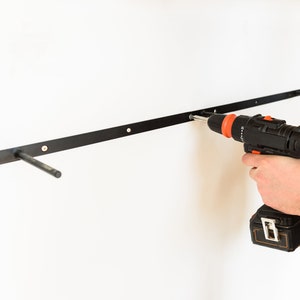 May include: A person uses a black and orange cordless drill to install a black metal shelf bracket on a white wall.