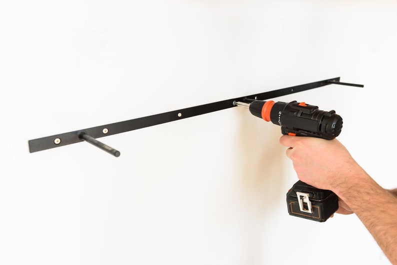 May include: A person uses a black and orange cordless drill to install a black metal shelf bracket on a white wall.