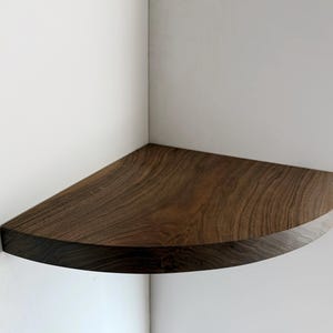 May include: A dark brown wooden corner shelf with a rounded edge. The shelf is mounted on a white wall.