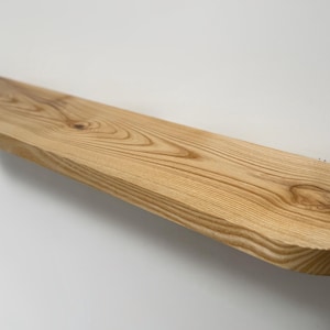 May include: A natural wood shelf with a rounded edge and a light brown finish. The shelf is mounted to a white wall.