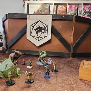 May include: A tabletop role-playing game scene with miniature figures, a wooden game box, and a banner with a dragon logo. The scene includes painted figurines on a brown surface, suggesting a fantasy or adventure theme.