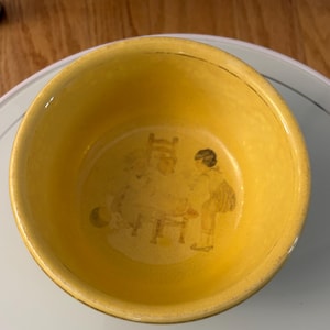 May include: A yellow ceramic bowl with a detailed illustration inside. The bowl has a glossy finish and a slightly irregular shape. The illustration depicts figures and furniture in a sepia tone. The bowl is sitting on a glass surface.