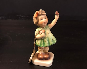 May include: A ceramic figurine of a young girl in a green dress, holding flowers and a small object. The figurine has a detailed face, brown hair, and is standing on a square base. A gold-colored charm hangs from a string.