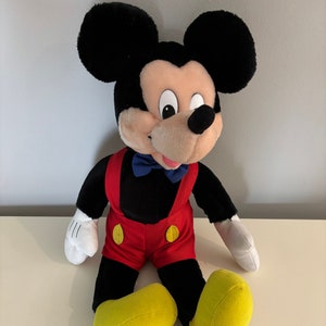 May include: A plush Mickey Mouse toy sitting with a red and black outfit, yellow shoes, and a blue bow tie.