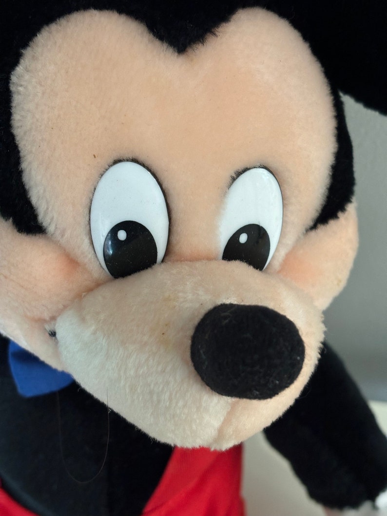 May include: A plush Mickey Mouse toy with black ears, a black nose, and white eyes. The toy is wearing a red and black outfit.