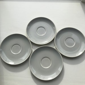 May include: Four white ceramic saucers with a slight gray tint. The saucers are arranged in a circle on a white surface.