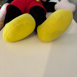 May include: A plush Mickey Mouse toy with black fur, red shorts, and yellow shoes. The toy is lying on its back with its arms outstretched.