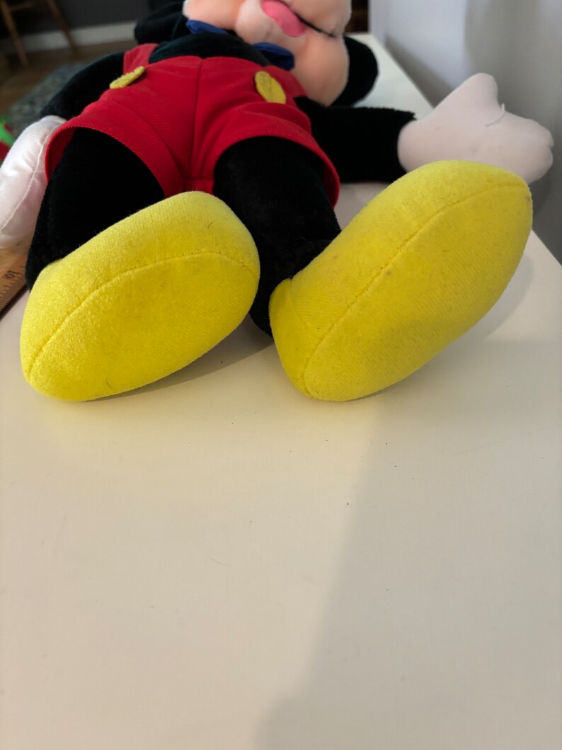 May include: A plush Mickey Mouse toy with black fur, red shorts, and yellow shoes. The toy is lying on its back with its arms outstretched.