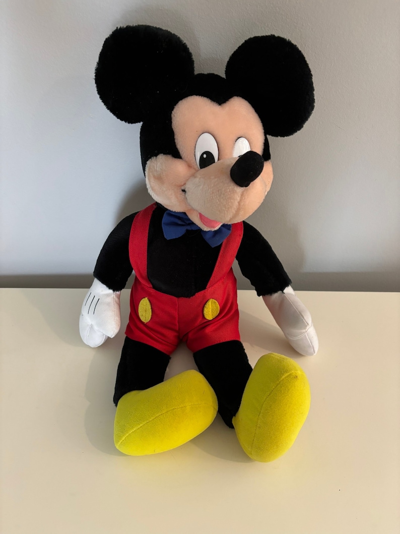 May include: A plush Mickey Mouse toy sitting with a red and black outfit, yellow shoes, and a blue bow tie.