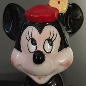 Pu&ograve; includere: Una statuetta in ceramica di Minnie Mouse con un cappello rosso con un fiore giallo. Ha orecchie nere, un naso nero e un viso bianco con ciglia nere. Sorride e indossa un vestito a pois rossi e bianchi.