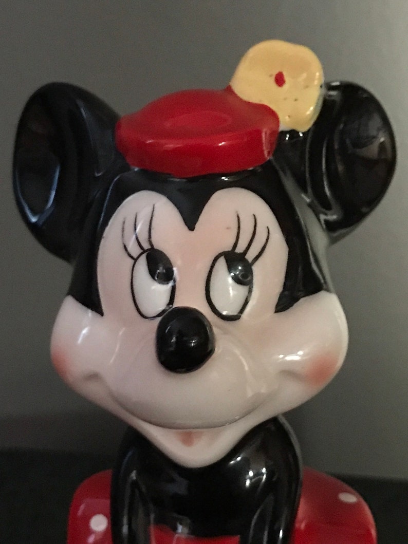 Pu&ograve; includere: Una statuetta in ceramica di Minnie Mouse con un cappello rosso con un fiore giallo. Ha orecchie nere, un naso nero e un viso bianco con ciglia nere. Sorride e indossa un vestito a pois rossi e bianchi.