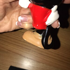 Pu&ograve; includere: Una statuetta di ceramica di Topolino, con una camicia rossa, pantaloncini neri e guanti bianchi. La statuetta &egrave; in piedi su una superficie di legno.