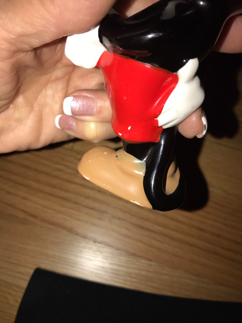 Pu&ograve; includere: Una statuetta di ceramica di Topolino, con una camicia rossa, pantaloncini neri e guanti bianchi. La statuetta &egrave; in piedi su una superficie di legno.