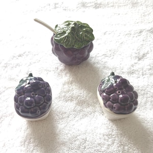 May include: A ceramic grape-shaped salt and pepper shaker set with a matching sugar bowl. The set features a purple color with green leaf accents. The sugar bowl includes a small spoon. The set is designed to resemble a bunch of grapes.