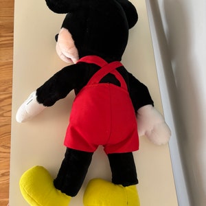 May include: A black plush Mickey Mouse toy wearing red overalls and yellow shoes. The toy is lying on its back with its arms outstretched.