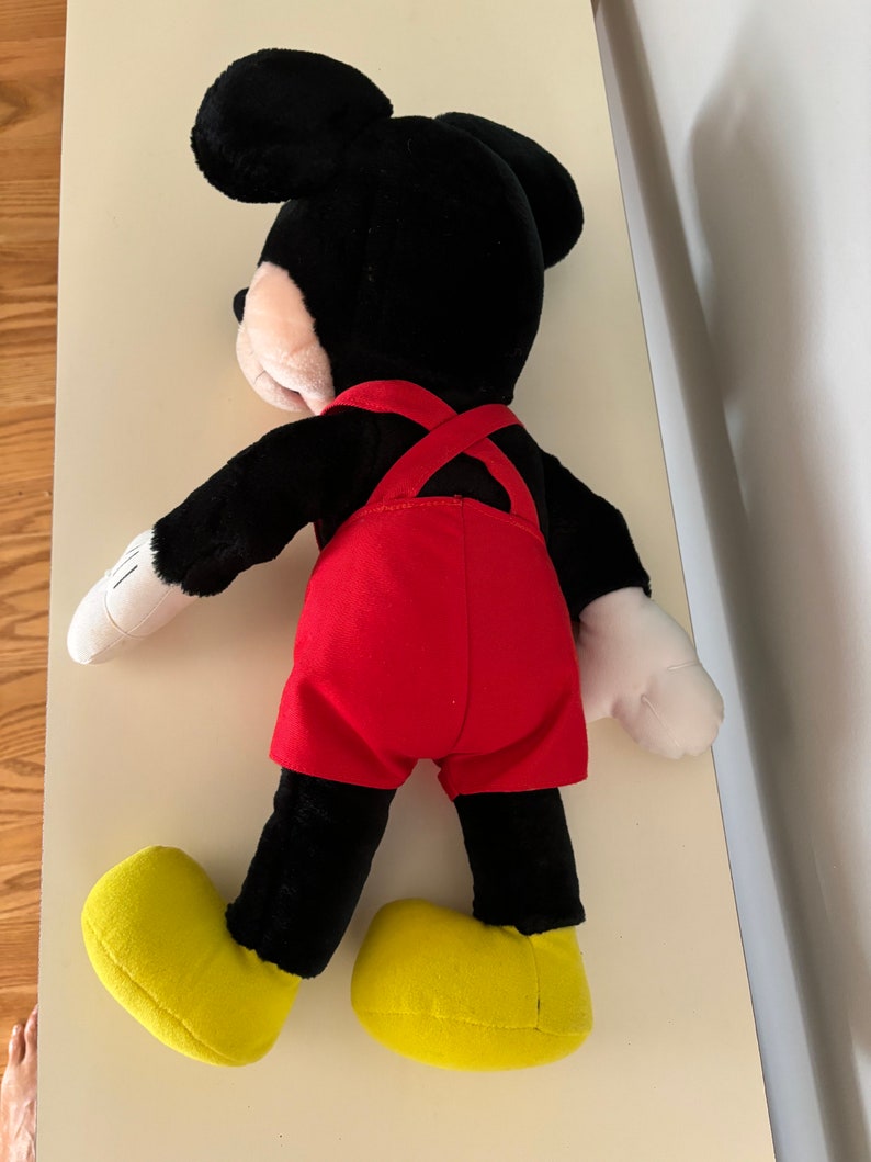 May include: A black plush Mickey Mouse toy wearing red overalls and yellow shoes. The toy is lying on its back with its arms outstretched.
