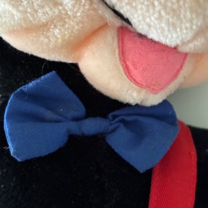 May include: A black plush toy with a white face and a pink tongue sticking out. The toy is wearing a blue bow tie and a red strap.