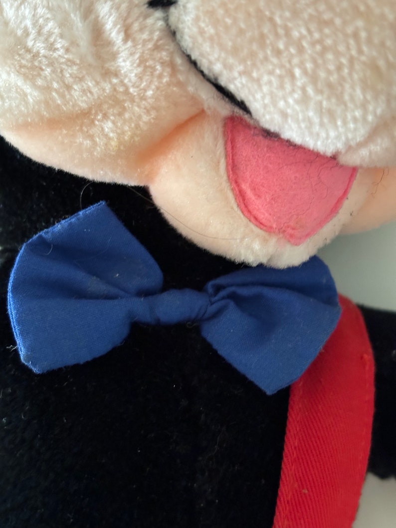 May include: A black plush toy with a white face and a pink tongue sticking out. The toy is wearing a blue bow tie and a red strap.