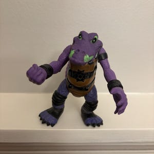 May include: A purple action figure of a humanoid alligator with green accents on its snout. The figure has black bands on its arms and legs, and a brown chest plate with a gear design. The figure is posed on a white shelf.