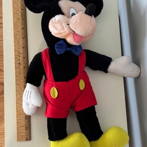 May include: A black and red plush Mickey Mouse toy with a blue bow tie. The toy is wearing red shorts with yellow buttons and has yellow shoes.
