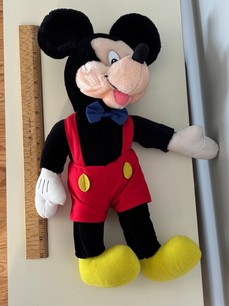 May include: A black and red plush Mickey Mouse toy with a blue bow tie. The toy is wearing red shorts with yellow buttons and has yellow shoes.