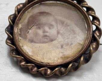May include: Vintage copper-coloured brooch featuring a circular photograph of a person. The photo is set within a round frame with a twisted rope pattern. The brooch has a pin fastening.