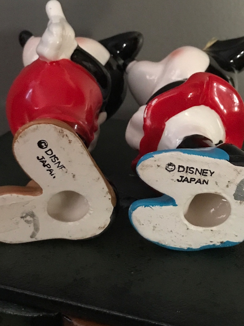Pu&ograve; includere: Un paio di figurine in ceramica di Topolino e Minnie Mouse. Entrambe le figurine indossano abiti rossi e bianchi. La parte inferiore di ogni figurina ha una base blu e bianca con il testo "DISNEY JAPAN" stampato sul fondo.
